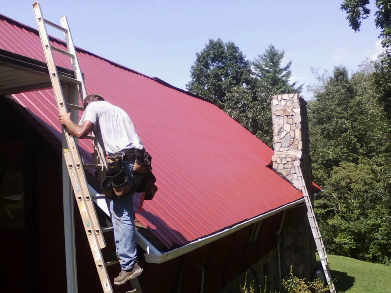 Roofer on a ladder beside a red metal roof and stone chimney — Soffit & Fascia Repair by Roofing Brothers in New Paris